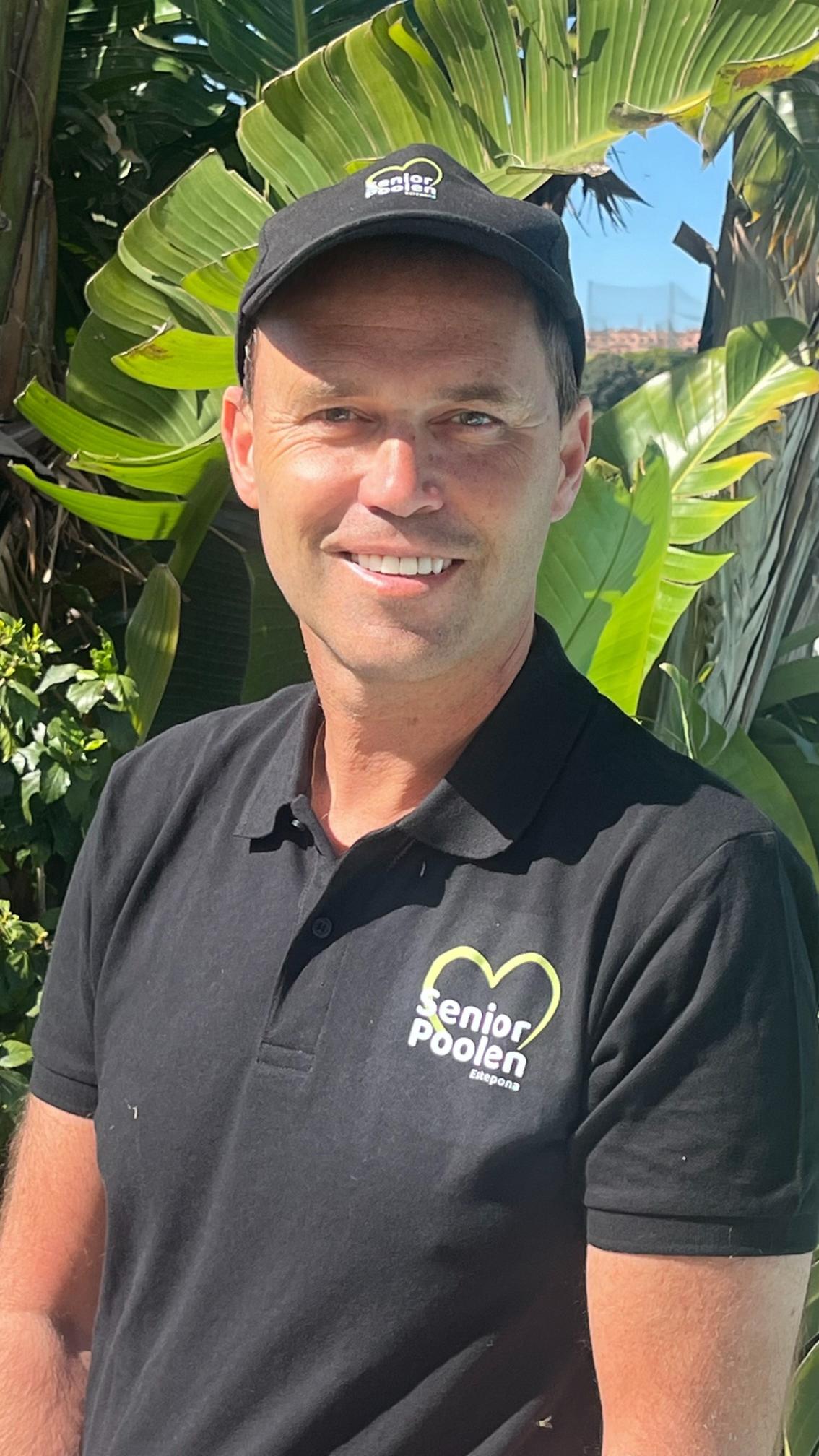 A man in a teal work jacket stands in a room with shelves in the background filled with various containers and supplies. He has short, tousled hair and a serious expression.